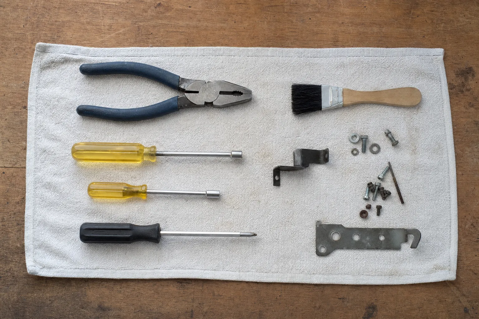Pliers, two screwdrivers and a parts brush laid out on a white cloth beside a hinge bracket, a long hinge plate and assorted screws and washers