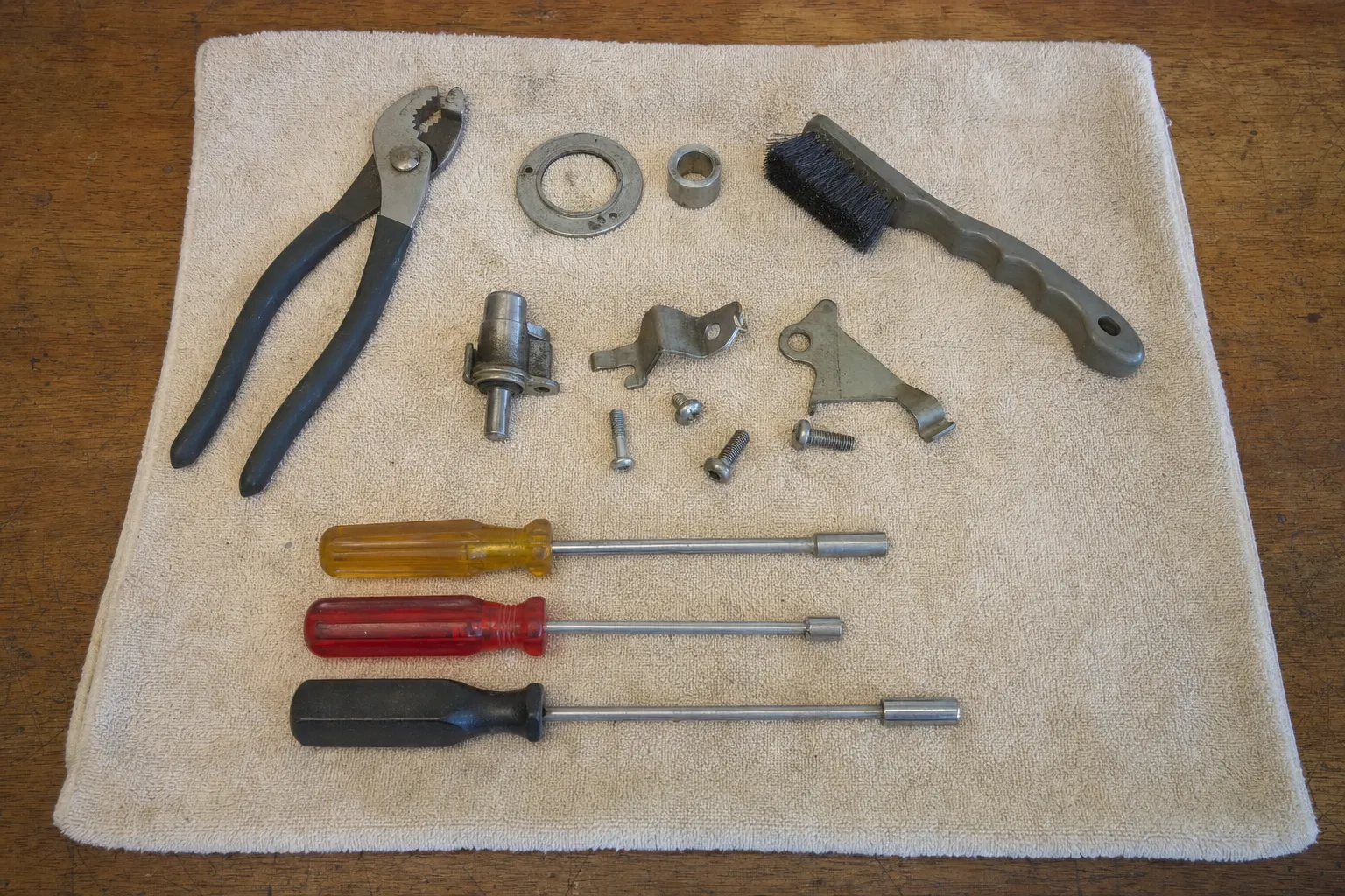 Slip-joint pliers, three nut drivers and a wire brush on a towel beside a ring plate, a bushing, a small cylindrical fitting on a bracket, two bent brackets and loose screws